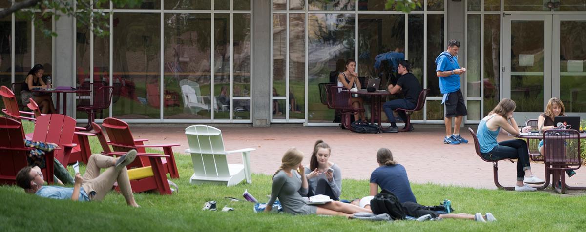 一群学生懒洋洋地躺在草地上，坐在玻璃墙建筑外的桌子旁
